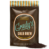 Front of Grady’s Cold Brew 16 oz coffee bag labeled “New Orleans-Style Coffee & Chicory.” The bag is brown with orange, white, and teal accents, and features the words “Cold Brew – Medium Dark Roast” in bold type. A colorful striped band at the top reads “Perfect for Hot and Cold Brewing.” Ground coffee is shown poured out in a pile in front of the bag.