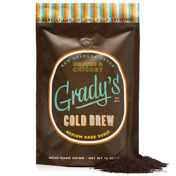 Front of Grady’s Cold Brew 16 oz coffee bag labeled “New Orleans-Style Coffee & Chicory.” The bag is brown with orange, white, and teal accents, and features the words “Cold Brew – Medium Dark Roast” in bold type. A colorful striped band at the top reads “Perfect for Hot and Cold Brewing.” Ground coffee is shown poured out in a pile in front of the bag.