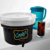 A clear-lidded Grady’s Cold Brew container with a black spout, filled with dark cold brew coffee. Positioned beside it are a translucent blue pitcher and a tall glass of iced coffee on a white surface.