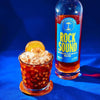 Glass of iced cold brew on a wooden coaster with ice cubes, garnished with a dried orange slice and banana chip, placed beside a bottle of Rock Sound Smoked Coconut Rum against a bright blue background.