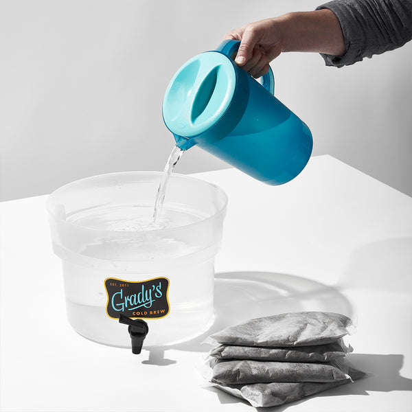 A hand pouring water from a blue pitcher into an empty Grady’s Cold Brew container, with a stack of large coffee bean bags placed on the table nearby.