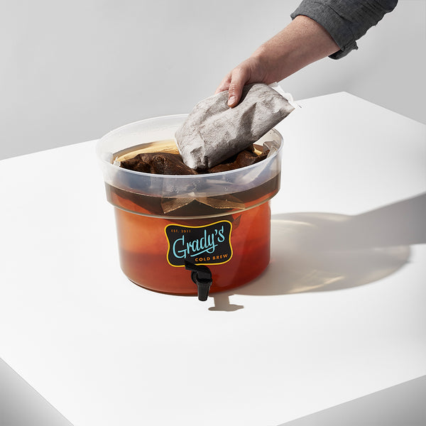 A hand placing a large cold brew bean bag into a Grady’s Cold Brew container, which is partially filled with steeping bags and water.