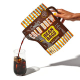 Hand pouring cold brew from a Grady’s Cold Brew Bean Bags “Spouch” into a glass filled with ice. The brown pouch features bold branding, a built-in handle, and colorful striped accents, with text indicating it makes 48 fluid ounces.