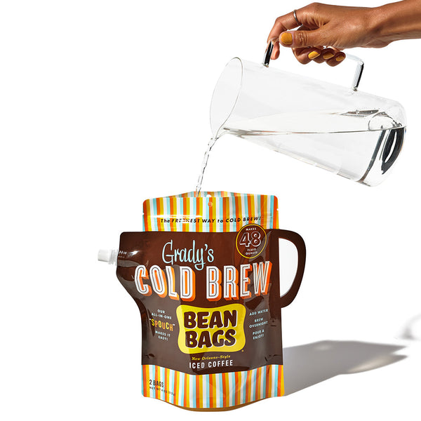Hand pouring water from a clear pitcher into a Grady’s Cold Brew Bean Bags “Spouch.” The brown pouch with a built-in handle and colorful striped top and bottom is labeled to make 48 fluid ounces of New Orleans–style iced coffee.