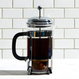 Glass French press coffee maker filled with freshly brewed dark coffee sitting on a white countertop. The French press features a black handle, chrome frame, and metal plunger lid. A white subway tile backsplash is visible in the background, creating a clean kitchen setting. The image highlights a simple brewing method for preparing hot coffee.