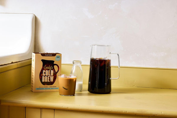 Lifestyle image of a yellow kitchen counter featuring a box of Grady’s Cold Brew ground coffee pouches, a glass of iced coffee with milk, a small milk bottle, and a clear pitcher of cold brew against a light wall background.