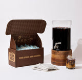 Open Grady's Cold Brew Bean Bags shipping box filled with reusable ice packs displayed beside a beverage dispenser filled with cold brew coffee. Three bean bags are placed in front of the box, and a small glass of iced coffee with cream sits next to the dispenser. The image showcases the complete brew-at-home experience, including the bean bags, cold brew dispenser, and finished coffee ready to serve.