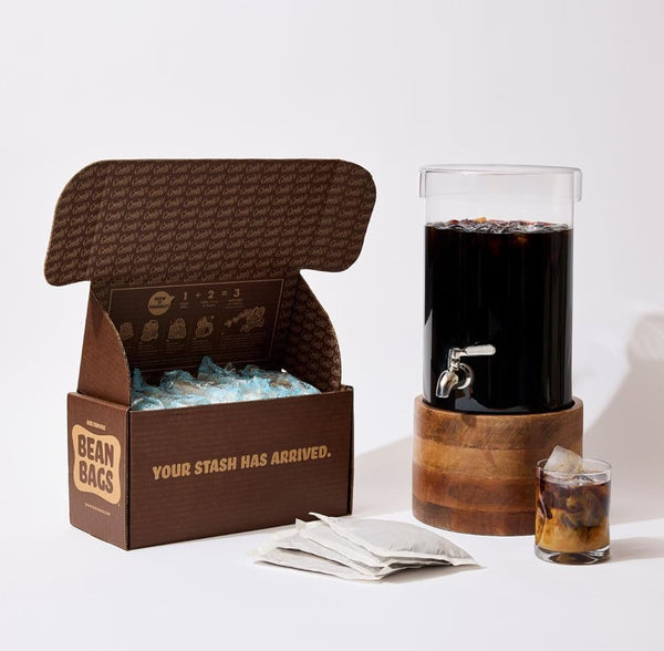 Open Grady's Cold Brew Bean Bags shipping box filled with reusable ice packs displayed beside a beverage dispenser filled with cold brew coffee. Three bean bags are placed in front of the box, and a small glass of iced coffee with cream sits next to the dispenser. The image showcases the complete brew-at-home experience, including the bean bags, cold brew dispenser, and finished coffee ready to serve.