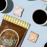 Overhead view of a bag of Grady's Cold Brew New Orleans-Style Coffee & Chicory ground coffee surrounded by cups of black coffee and powdered sugar-covered beignets on a light blue background. The packaging highlights that the coffee is suitable for both hot and cold brewing. The image evokes a classic New Orleans coffee-and-beignet pairing and showcases the coffee's connection to traditional coffee and chicory flavors.