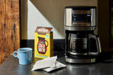 Lifestyle image of a kitchen counter with a drip coffee maker, a box of Grady’s Cold Brew ground coffee, a blue coffee mug, and individual coffee bean bags laid out in front of the box.