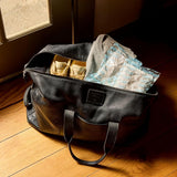 Black leather tote bag sitting on a hardwood floor near a glass door, partially packed with casual clothing, shoes, and several Grady's Cold Brew ice packs. Warm sunlight streams into the room, illuminating the bag and its contents. The image suggests using Grady's reusable ice packs to help keep items cool while traveling or on the go.