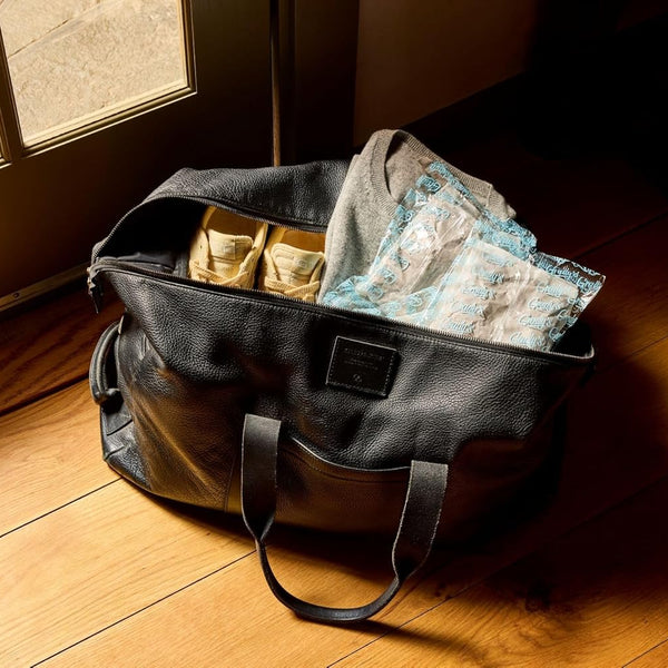 Black leather tote bag sitting on a hardwood floor near a glass door, partially packed with casual clothing, shoes, and several Grady's Cold Brew ice packs. Warm sunlight streams into the room, illuminating the bag and its contents. The image suggests using Grady's reusable ice packs to help keep items cool while traveling or on the go.