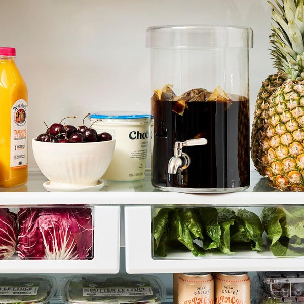 Inside a refrigerator, a glass beverage dispenser filled with cold brew coffee and ice sits on a shelf alongside fresh fruit and groceries. Oranges, cherries, pineapple, juice, yogurt, leafy greens, and berries surround the dispenser. The image highlights cold brew coffee being stored in the refrigerator and staying fresh alongside everyday household foods and beverages.