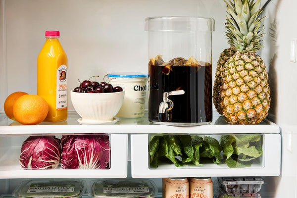 Interior view of a refrigerator showing a glass container filled with cold brew coffee and ice, placed on a shelf with oranges, juice, cherries, yogurt, lettuce, and a pineapple.