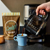 Freshly brewed coffee being poured from a drip coffee maker into a light blue mug on a kitchen countertop. A bag of Grady's Cold Brew New Orleans-Style Coffee & Chicory medium dark roast ground coffee stands beside the mug, with a small cream pitcher nearby. The scene demonstrates the coffee being prepared hot, highlighting that the grounds can be used for both hot and cold brewing. Warm natural light illuminates the kitchen setting.