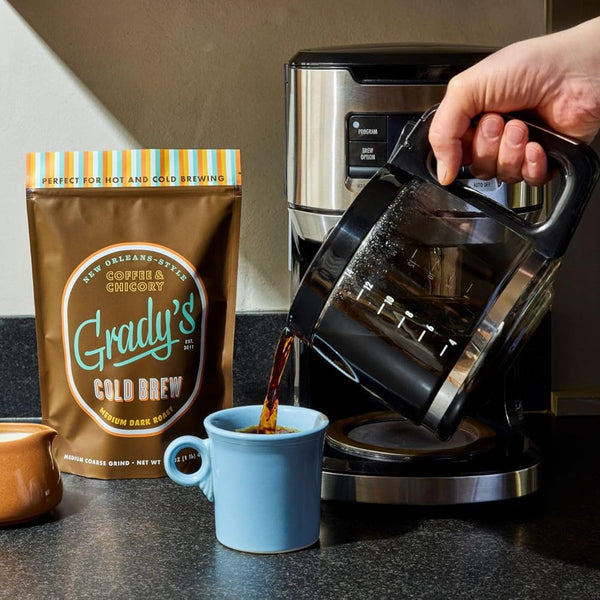 Freshly brewed coffee being poured from a drip coffee maker into a light blue mug on a kitchen countertop. A bag of Grady's Cold Brew New Orleans-Style Coffee & Chicory medium dark roast ground coffee stands beside the mug, with a small cream pitcher nearby. The scene demonstrates the coffee being prepared hot, highlighting that the grounds can be used for both hot and cold brewing. Warm natural light illuminates the kitchen setting.