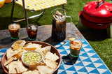 Sunny outdoor scene showing a wooden table with a pitcher of iced cold brew, multiple iced coffee drinks, and a bowl of tortilla chips with guacamole on a patterned tablecloth.
