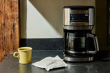Lifestyle image showing a drip coffee maker on a kitchen counter next to a yellow mug of coffee and a single-use coffee brewing pouch.