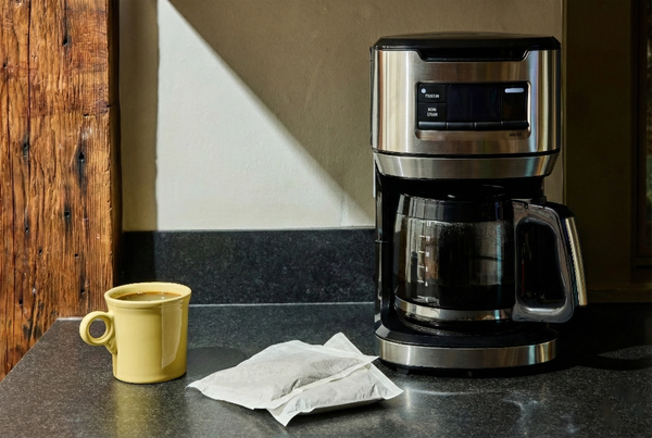 Lifestyle image showing a drip coffee maker on a kitchen counter next to a yellow mug of coffee and a single-use coffee brewing pouch.